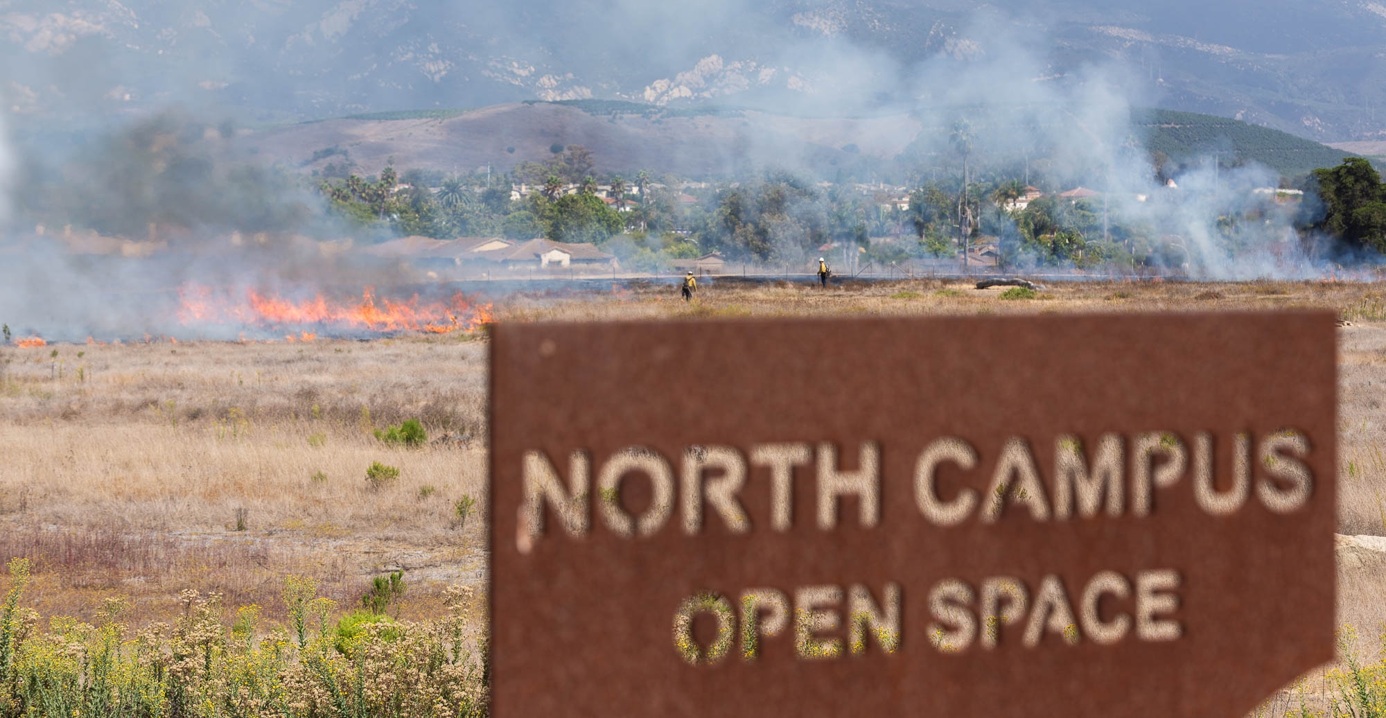 A sign reading "North Campus Open Space" in the foreground with a smoking brush fire in the background