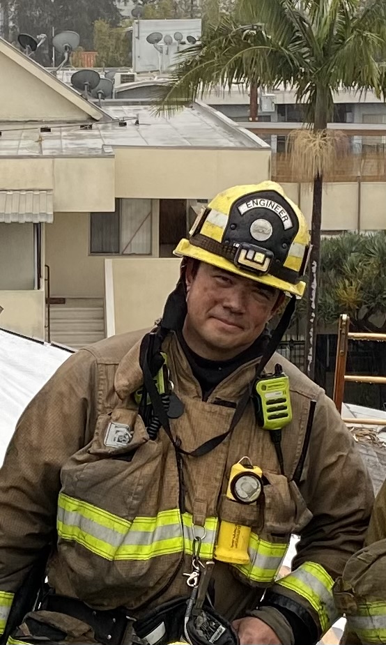 Derek Urwin smiles at the camera wearing firefighting gear: a yellow helmet and a soot-covered heavy jacket.