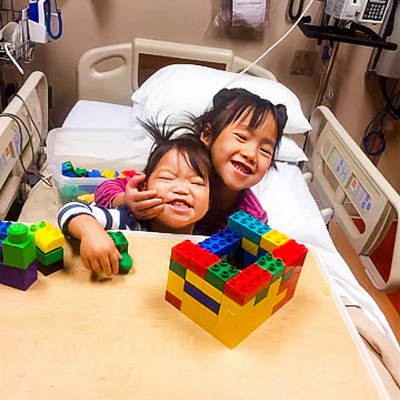 Ada and Lily smile in a hospital bed, playing with Legos