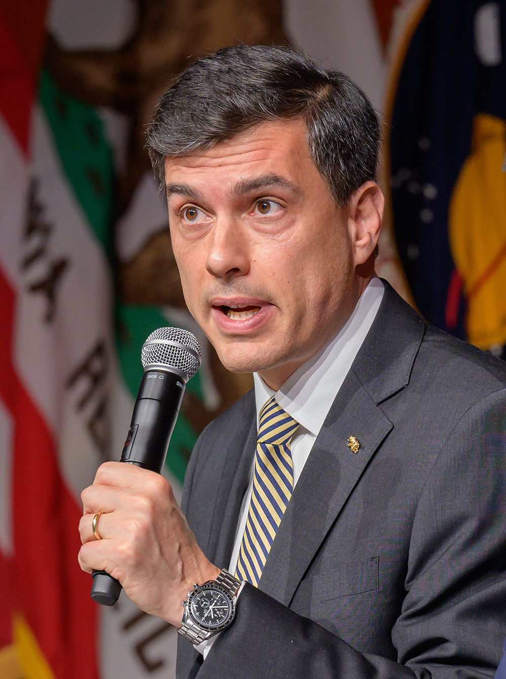 A white man in a suit holds a mic with the California flag in the background