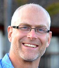 Trent Biggs, with close-cropped hair and a trim gray beard and glasses, smiles at the camera in a close-up image