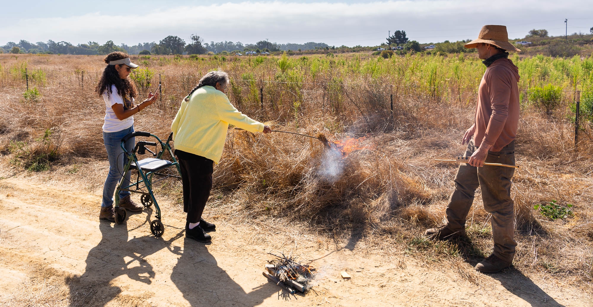 An elder woman in a yellow sweater stands near a mobility device to light a fire in dry brush, watched by two people.