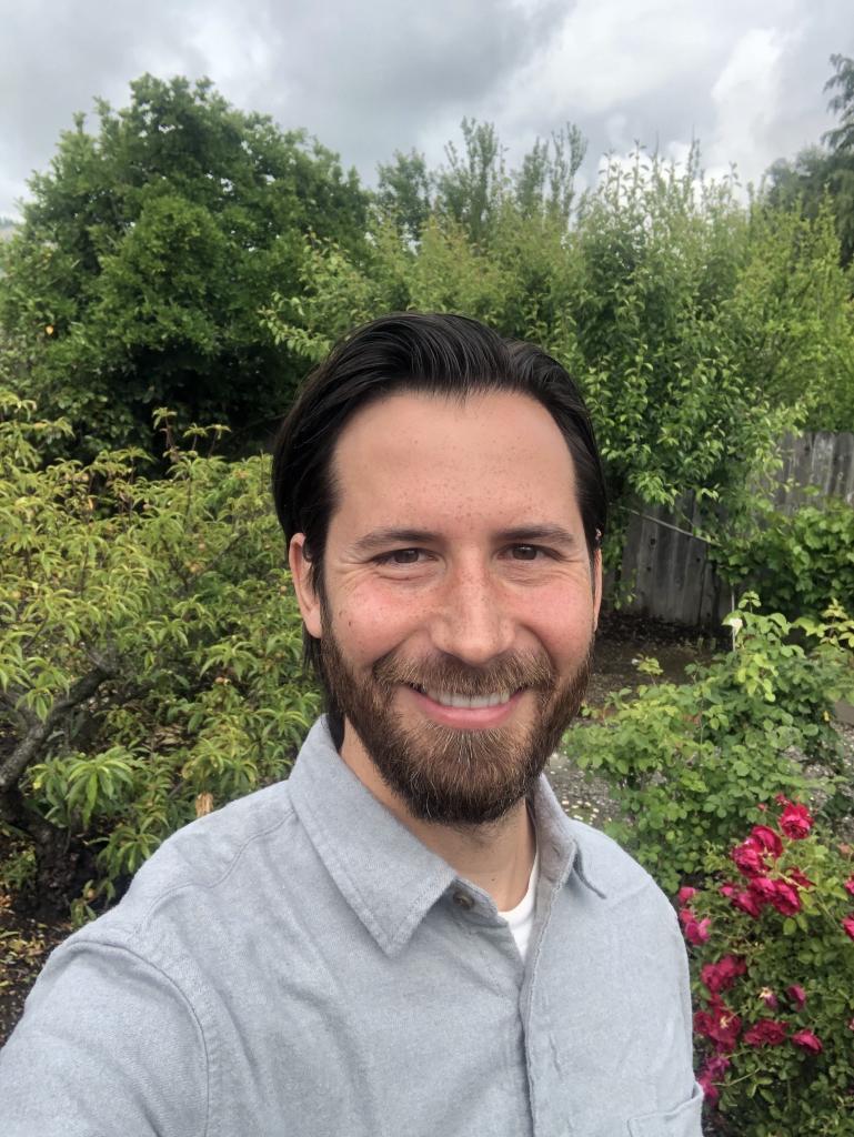 Peter Nelson, bearded wearing a gray shirt, smiles at the camera in a shoulders-up selfie, with greenery in the background