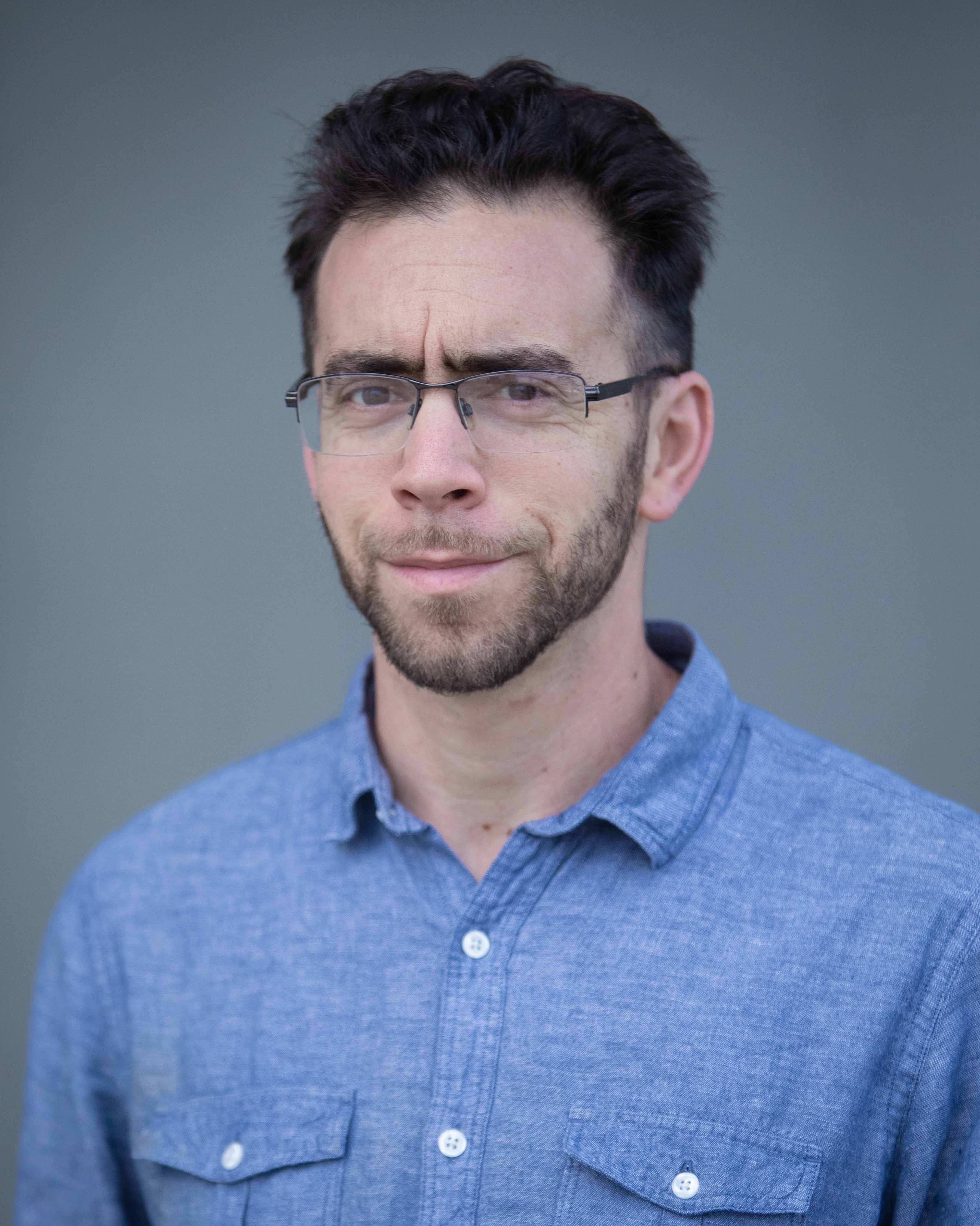 Tony Marks-Block, close-cropped dark beard and dark hair with glasses, wearing a blue shirt, in a shoulders-up portrait against a gray background