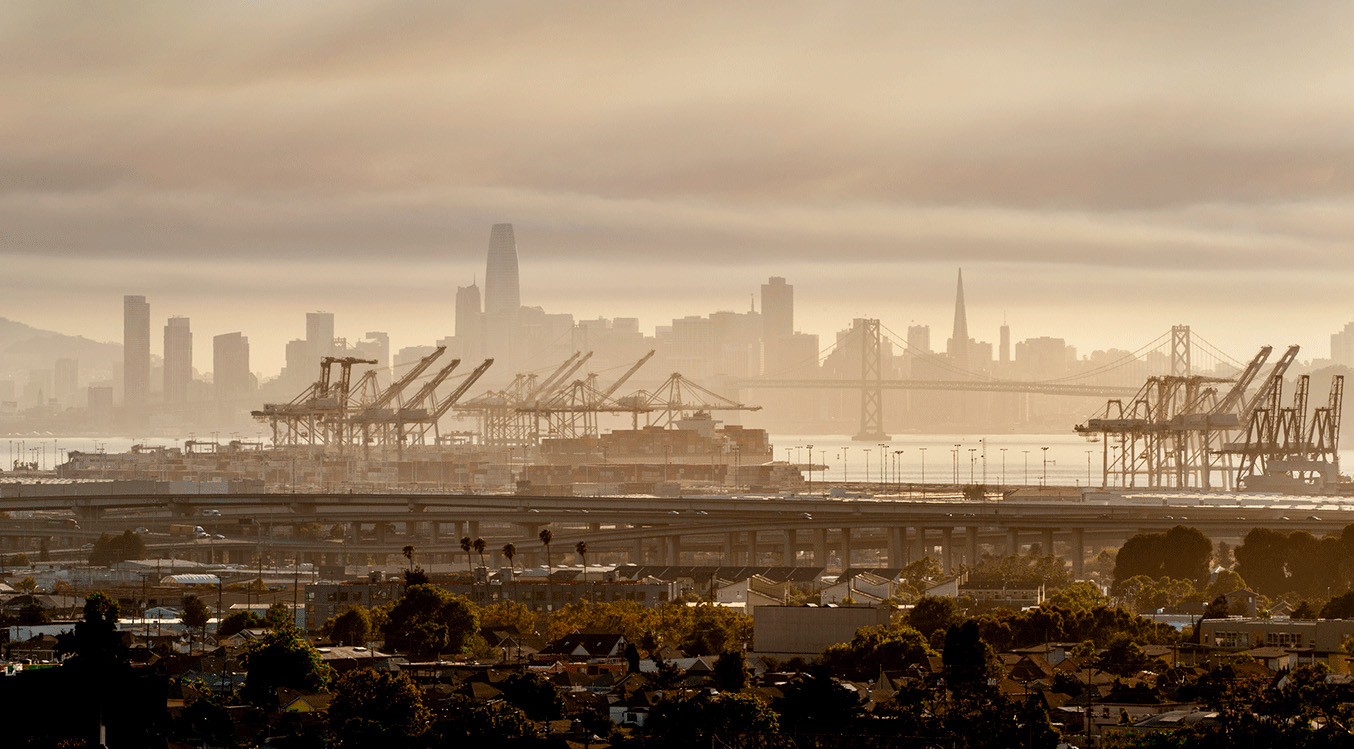 Skyline of San Francisco in the background, cranes in the Port of Oakland in the middle, and West Oakland in the near ground, under a cloudy/hazy sky
