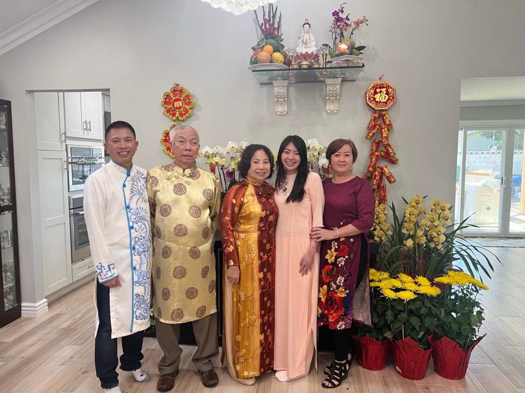 Chelsey Nguyen, pictured with her family, including her grandfather, wearing colorful clothing, posing in front of a collection of potted plants.