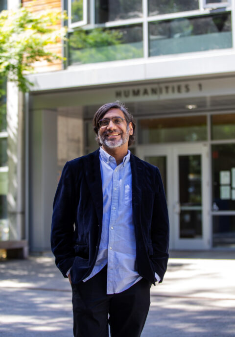 Pranav Anand, wearing blue button up, dark jacket, smiles at the camera in a knees-up portrait in front of a building labeled "HUMANITIES 1"