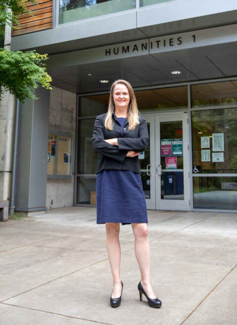 A blonde woman wearing a blue dress and black jacket smiles with arms folded across her chest standing in front of a building labeled "HUMANITIES 1"