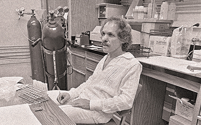Arnold Mandell in a candid photo at a desk in a lab. The photo and Mandell's fashions (shaggy hair, mustache, deep v-neck top) have a 1970s vibe.