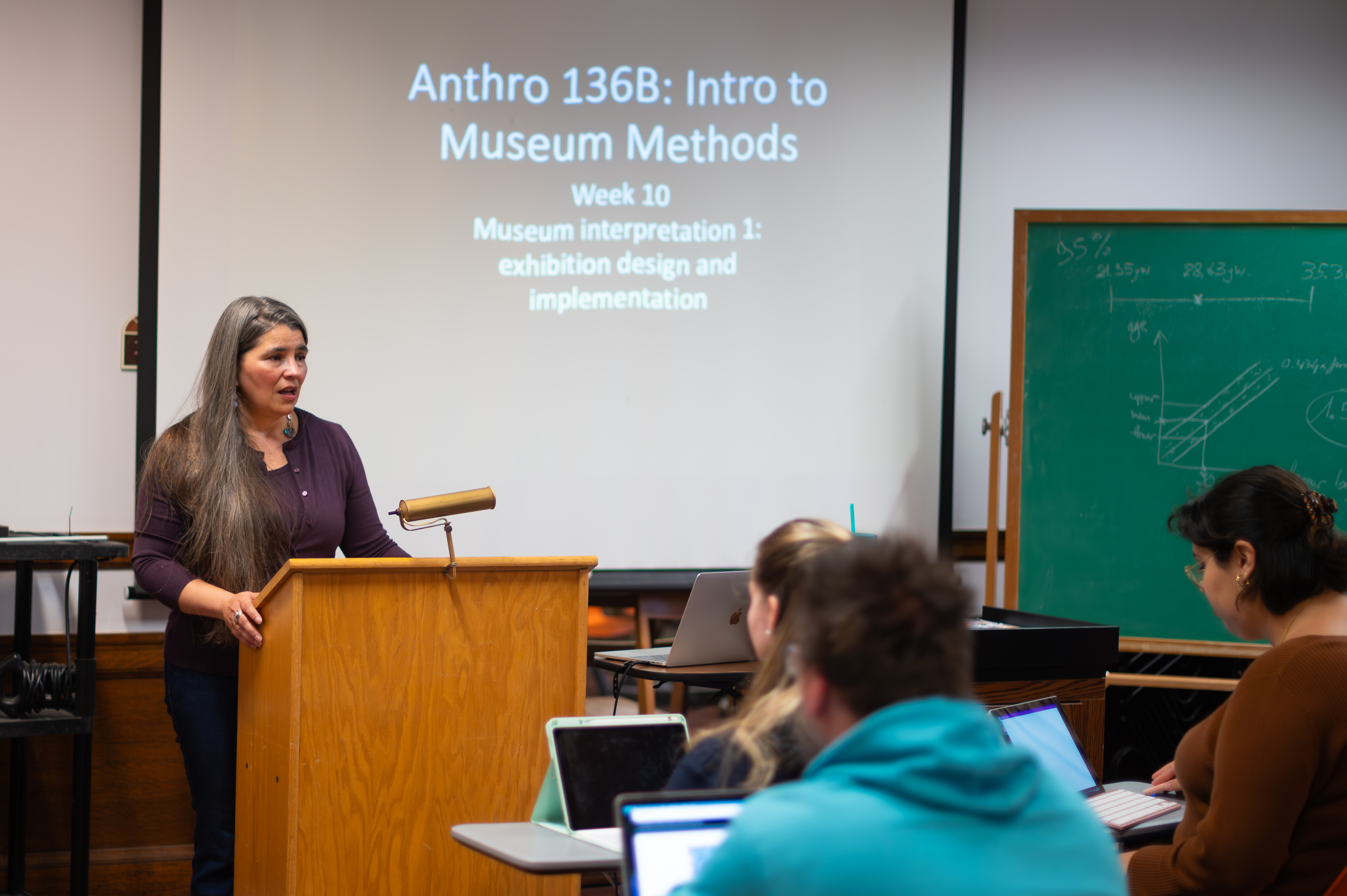 Carolyn Smith speaks at a lectern with a screen projection behind her reading "Anthro 136B: Intro to Museum Methods". Students at desks listen and take notes.