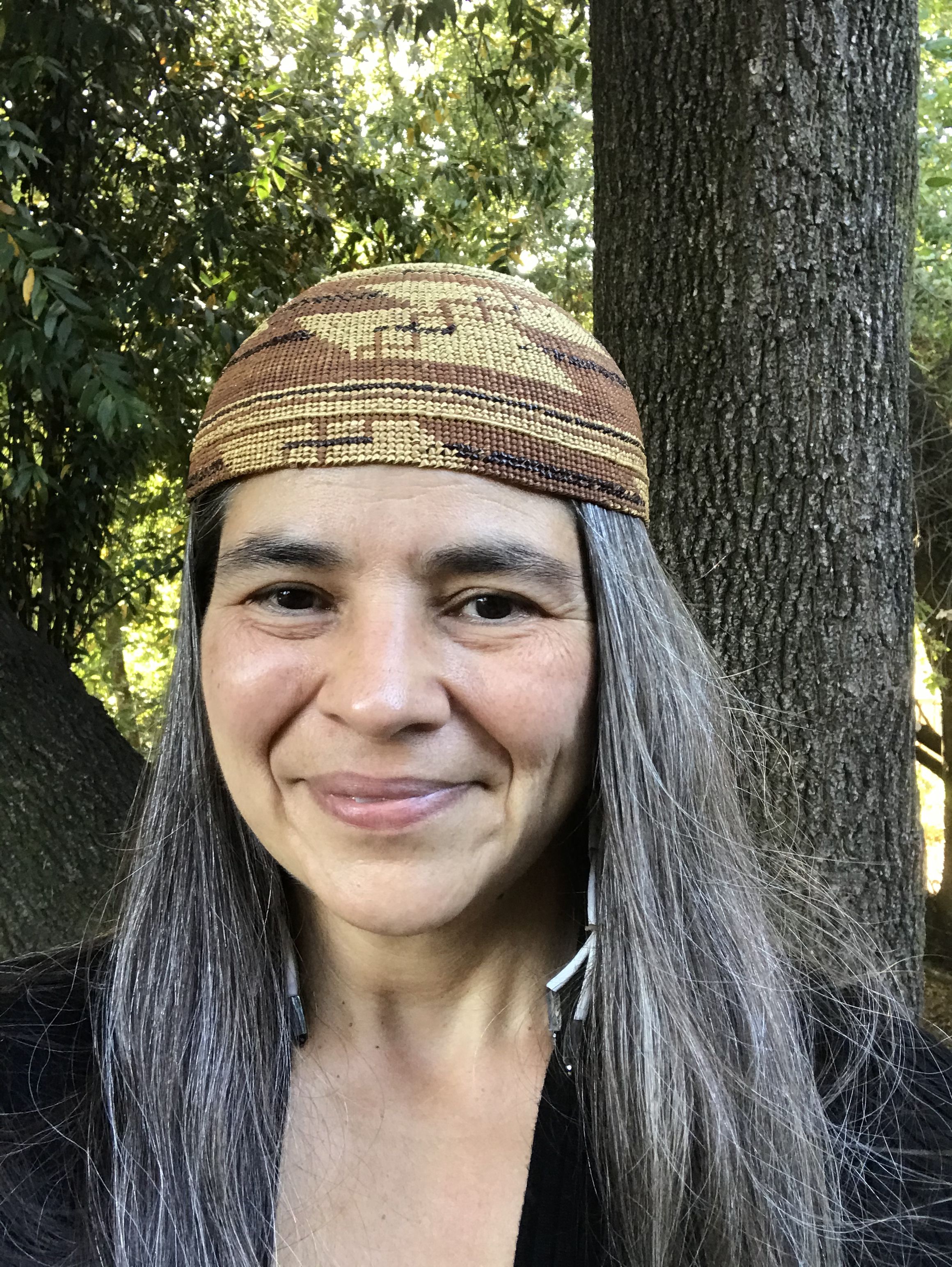 Carolyn Smith smiles at the camera, wearing a woven basket on her head. A tree trunk is in the background.