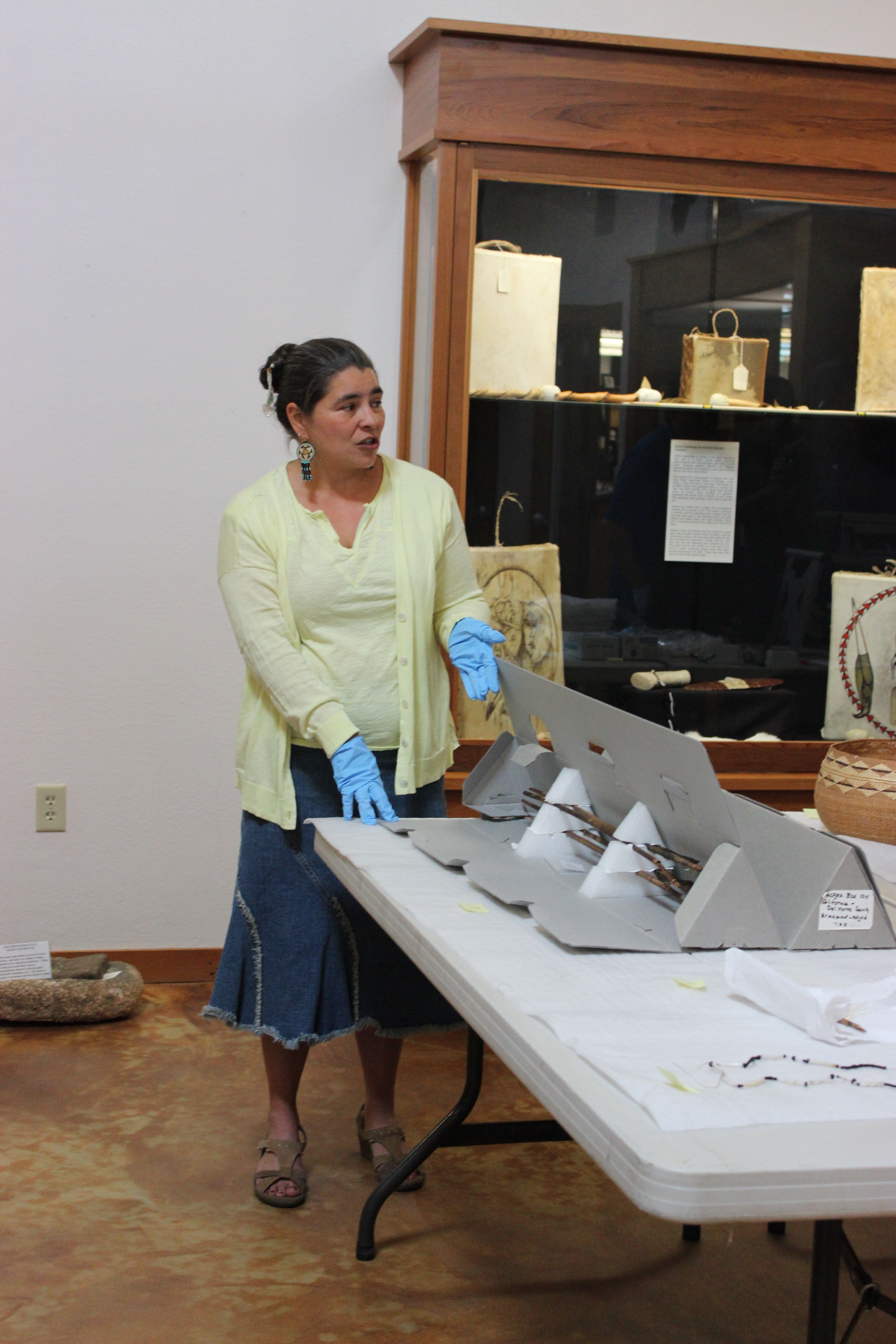 Carolyn Smith wears blue rubber gloves and talks to a person standing out of frame, standing in front of a wooden display shelf and in front of a table holding artifacts