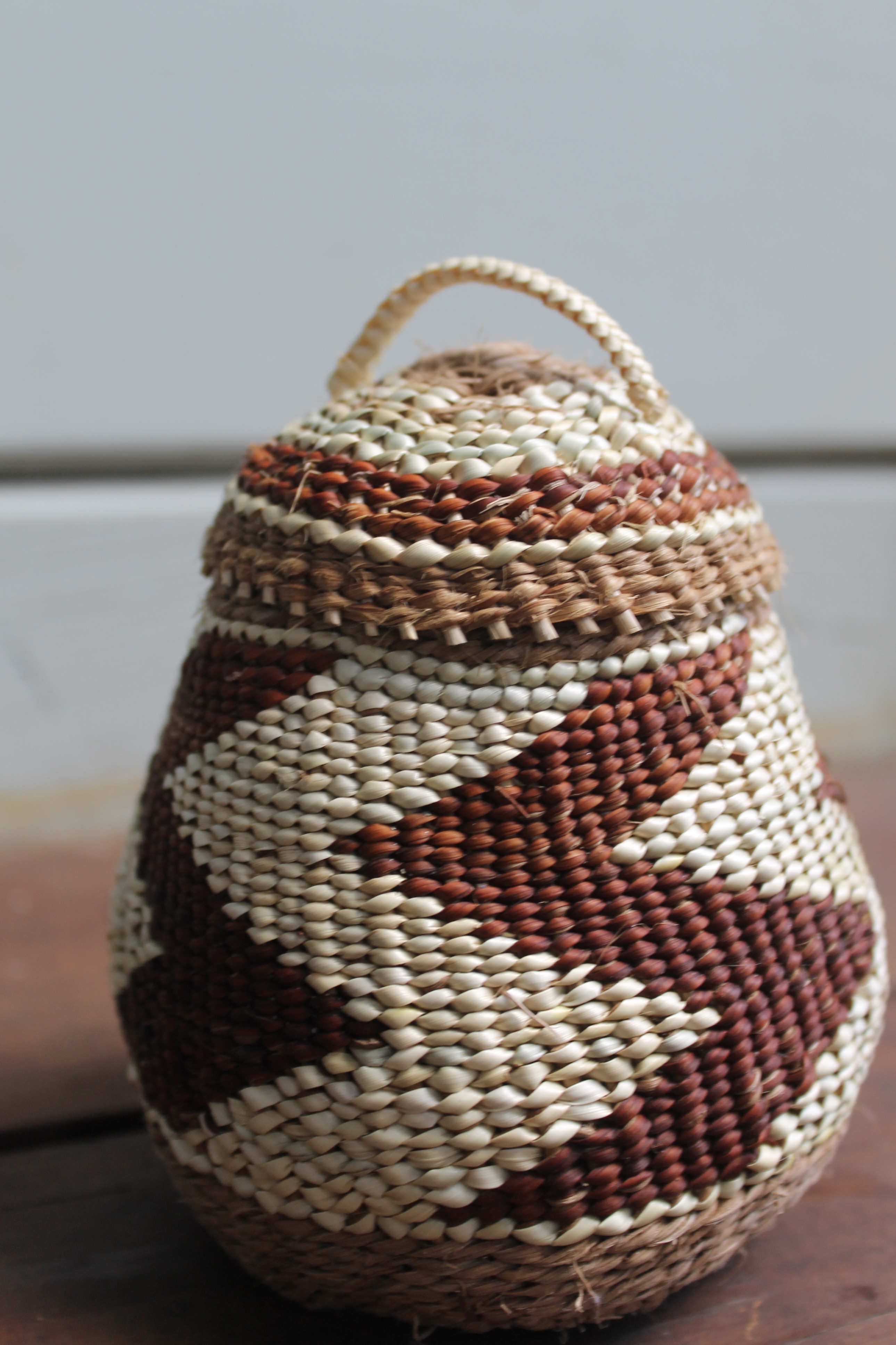 A cone-shaped brown and white woven basket with a zig zag pattern and a lid with a handle on a wooden shelf