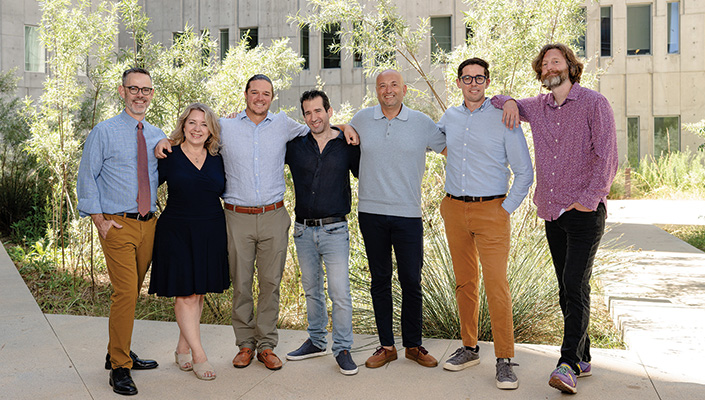 Seven adults in business casual attire with their arms around each others' shoulders smile for a picture in a courtyard with trees and a building in the background.