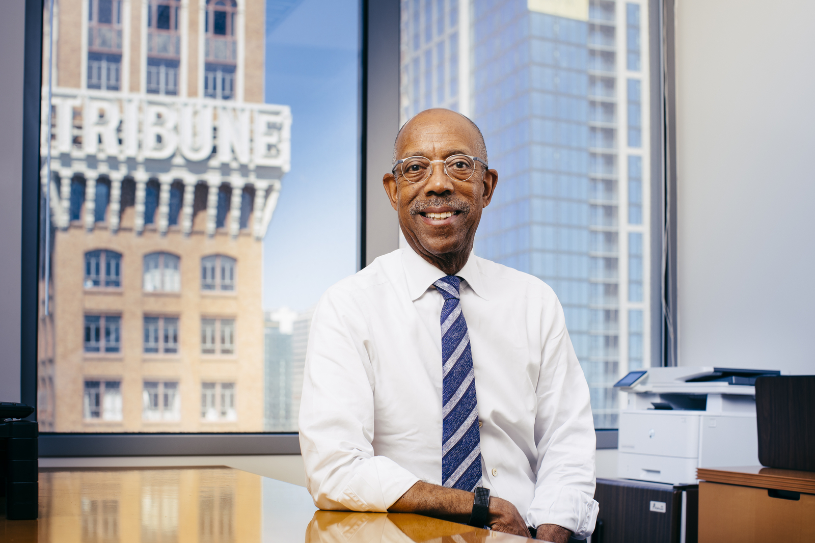 UC President Michael V. Drake, M.D., smiles for a portrait while sitting at a desk in his office. The Tribune Tower is visible out the window behind him. 