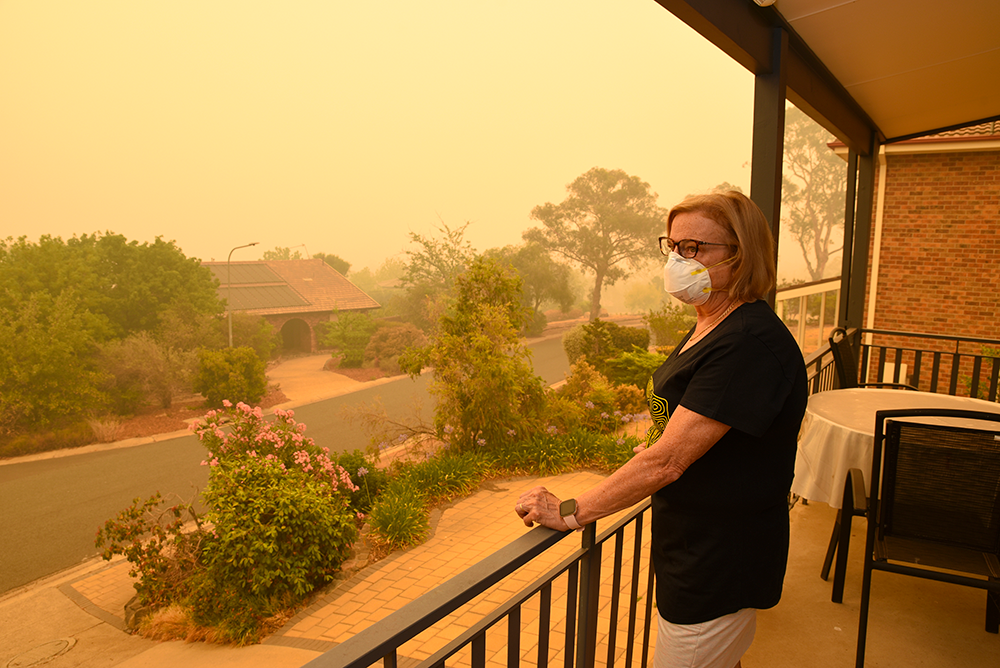 A woman wearing a mask stand on a balcony looking over a residential street. Smoke turns the air hazy and orange.