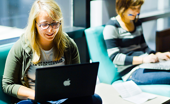 White student with blonde hair and glasses typing at computer another young woman typing behind