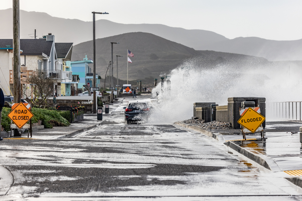 A car gets swamped by a big wave in Pacifica, California. Road signs warn of flooding.