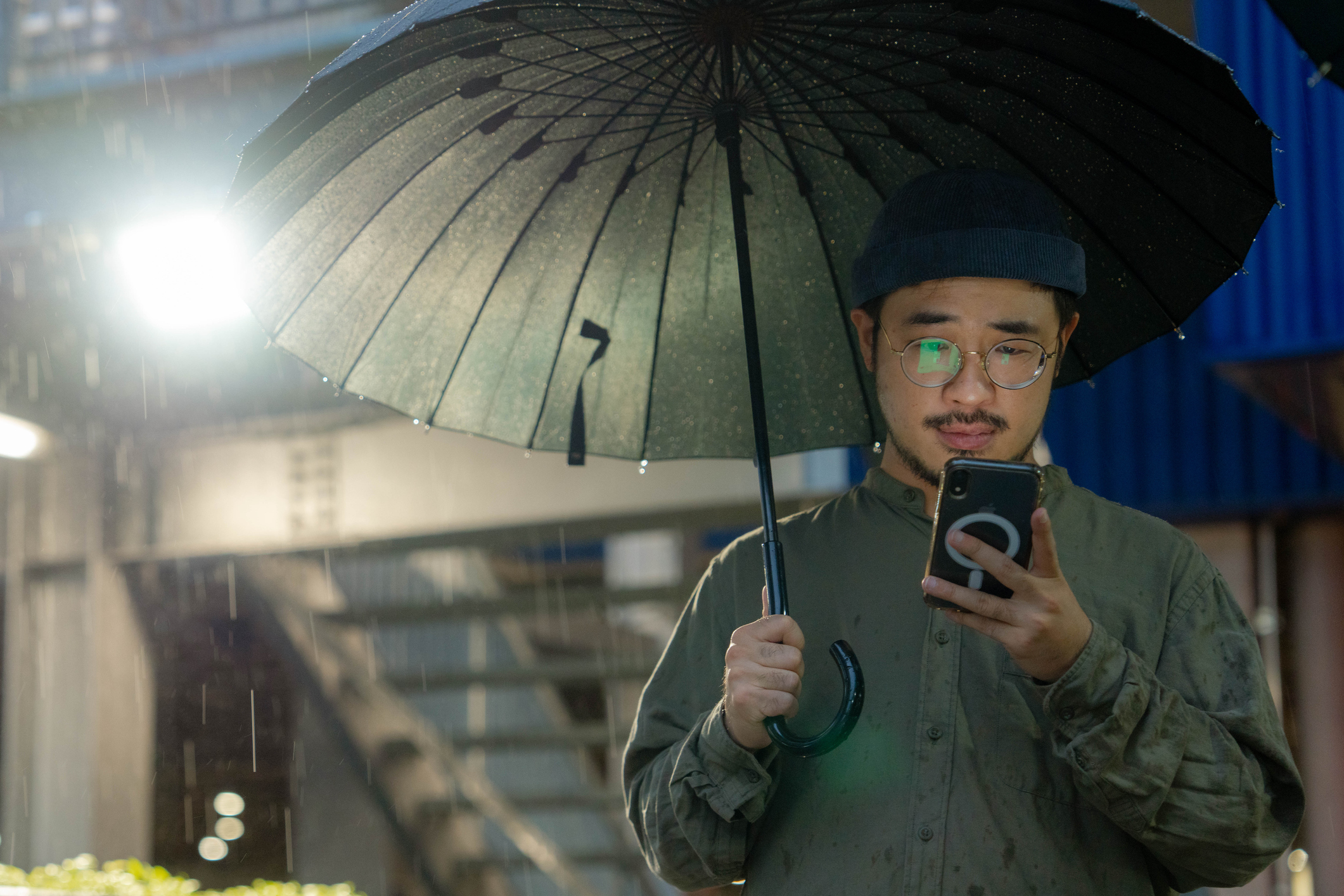 A man wearing a beanie and glasses looks at his smartphone under an umbrella with an annoyed expression. A floodlight appears over his shoulder.