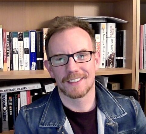 A white man with red hair and beard and dark rimmed glasses smiles at the camera in a shoulders-up picture, wearing a jean jacket