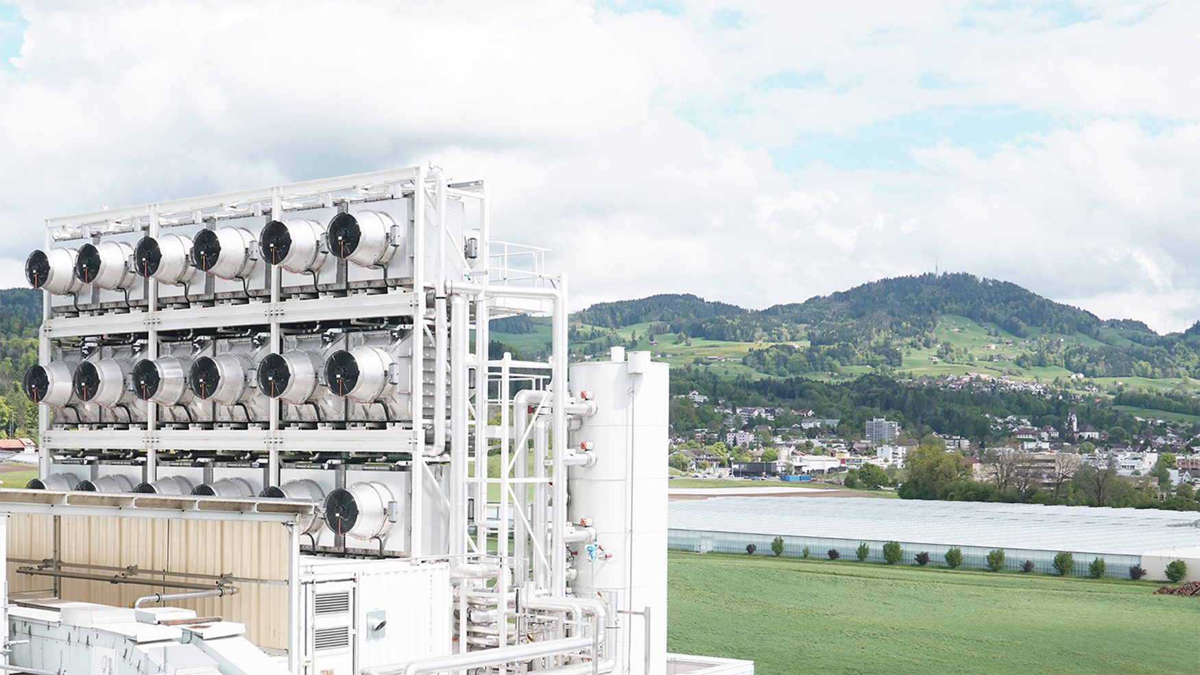 Photograph of a big white industrial facility with silver fans on the banks of a river with green hills in the background and a blue sky with puffy clouds