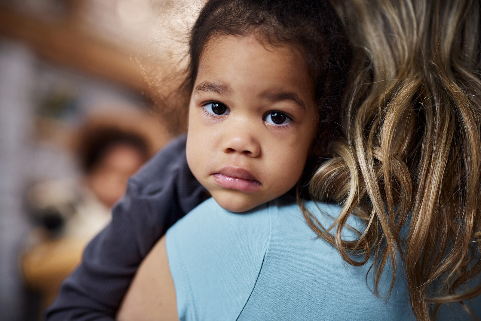 Toddler looks soberly at the camera, held over the shoulder of an adult with long blonde hair, whose back is to the camera