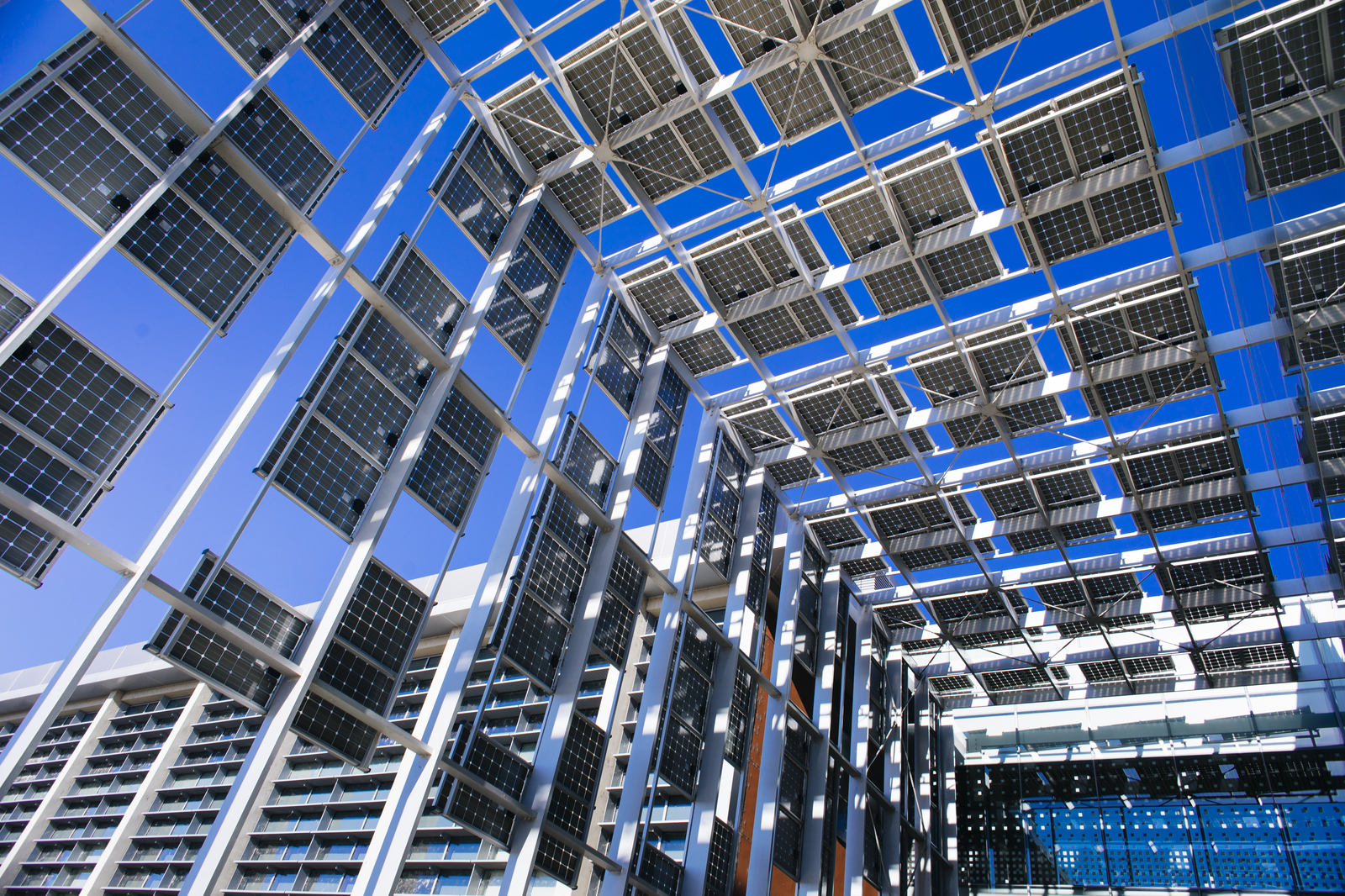 Looking up at a tall breezeway leading into a glass-walled building - the breezeway is constructed of solar panels on a scaffold. The sky is very blue.