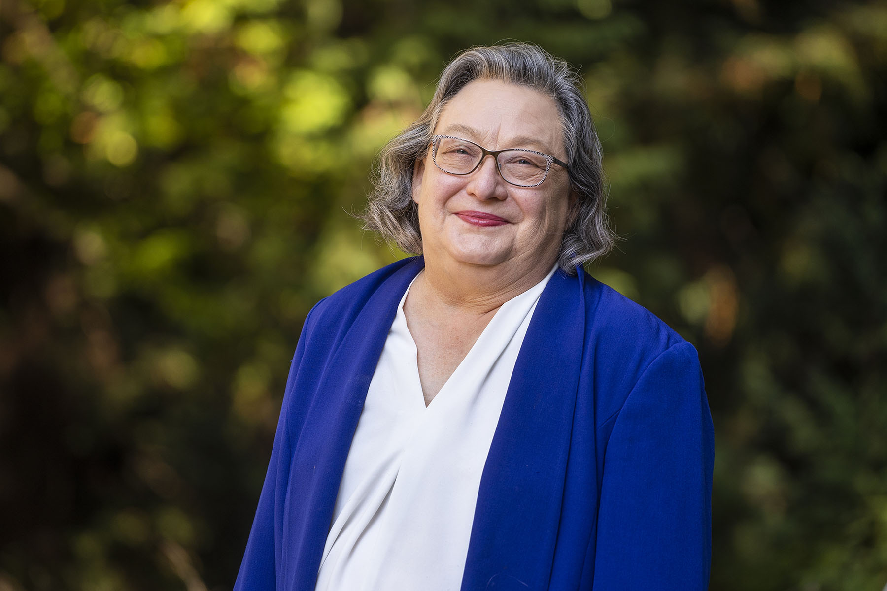 Portrait of Chancellor Larive, who smiles at the camera wearing a blue blazer and white blouse, against a backdrop of greenery.