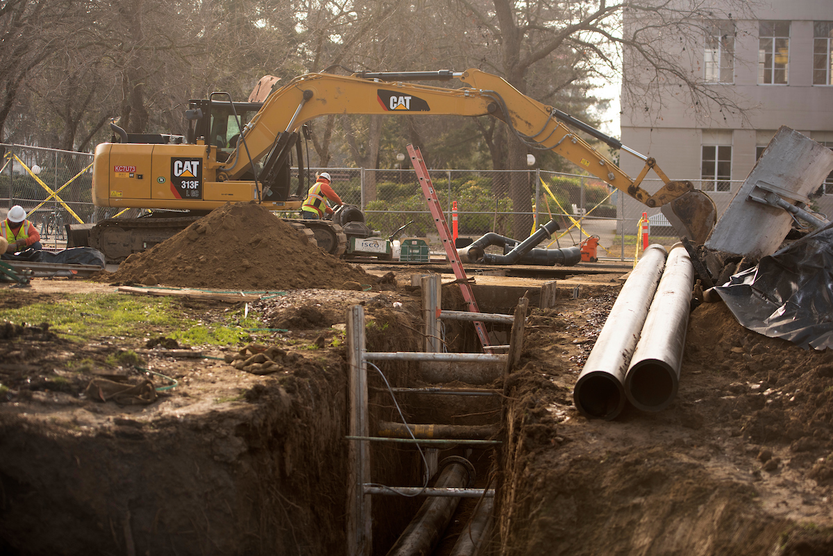 A person in construction safety gear operates a large yellow excavator above a deep trench with pipes laying parallel.