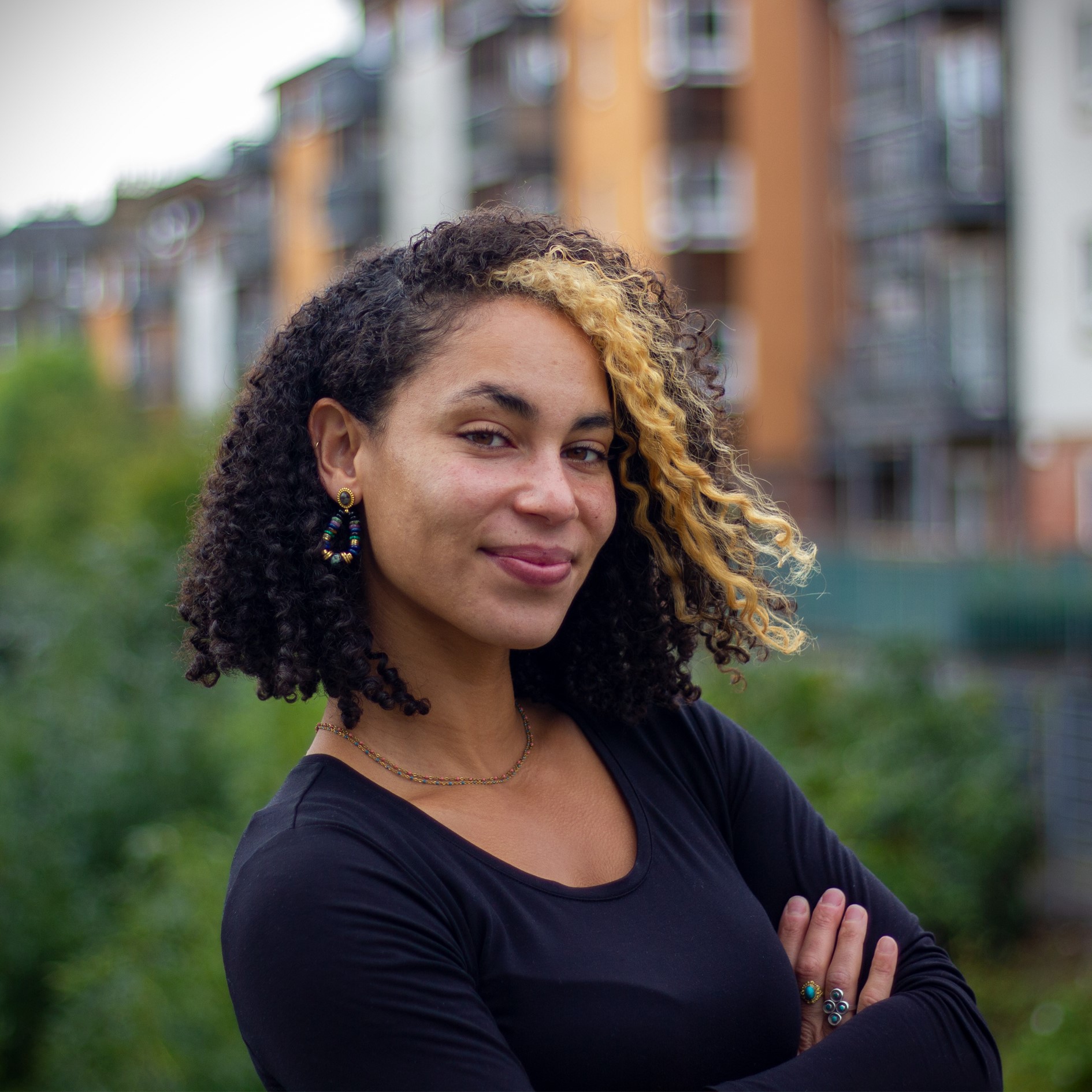 Laura Simone Lewis smiles for a headshot, arms crossed, wearing a black top. Apartment buildings are in the backdrop.