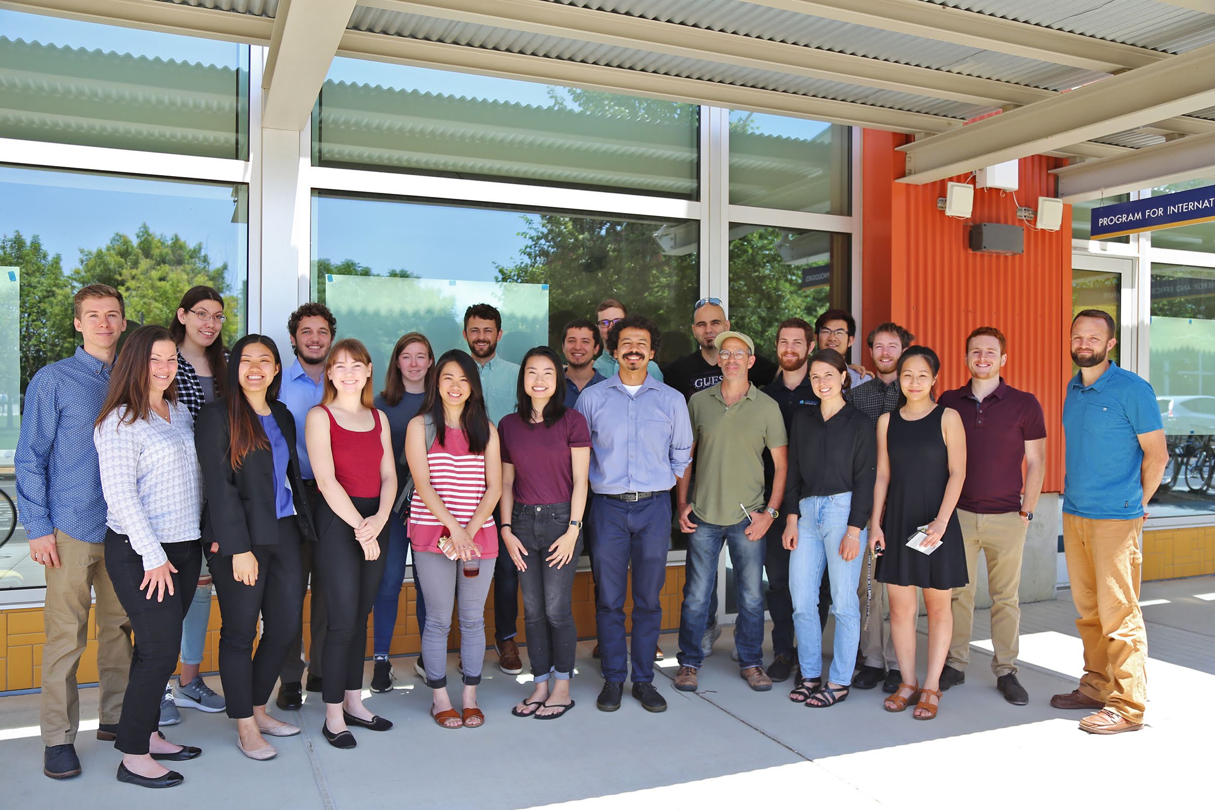 A posed group portrait of ~20 people in business casual attire standing in front of a building with big windows.