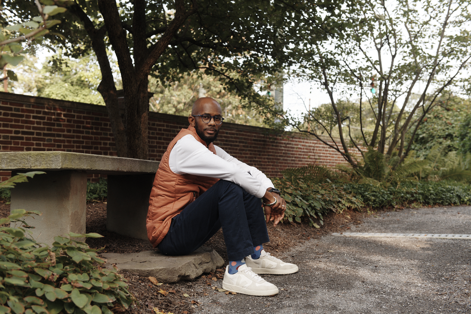 Young Black man in glasses and an orange vest sitting at the base of a bench on the Harvard campus