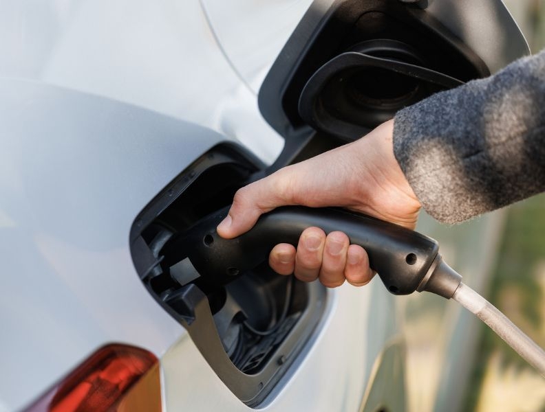 A hand holds a plug in the charging port of a white electric vehicle