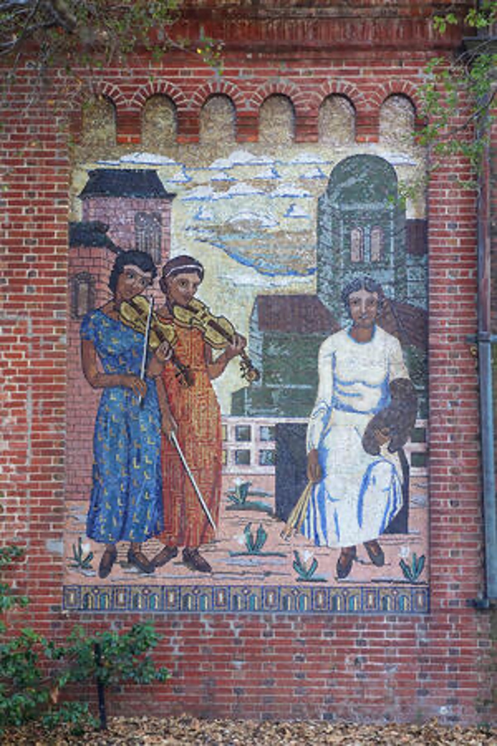 Tile mosaic showing a man in a white robe being played music by two women, one in a blue dress and one in an orange dress, on a brick wall.
