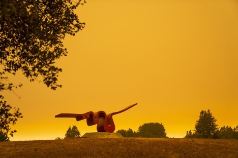 The Porter Squiggle, a large red sculpture, with tree foliage in the foreground. The sky is orange with wildfire smoke.