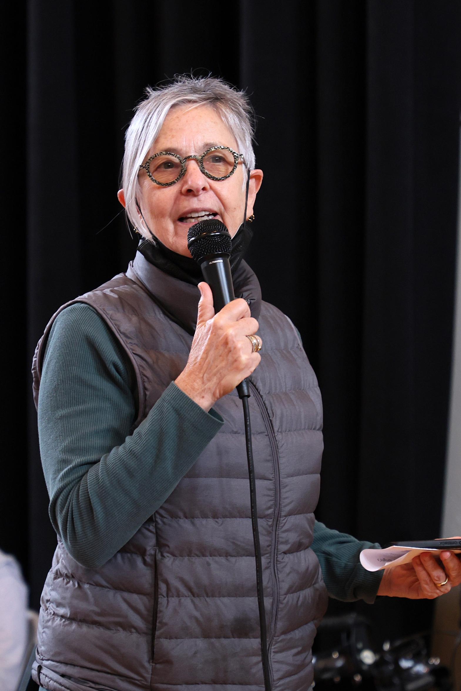 Victoria Marks, short gray hair and glasses, speaks into a microphone, against a black backdrop, wearing gray puffy vest and green long sleeve shirt.