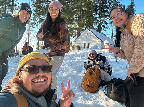 Students in a snowy forest setting make the victory sign, with a cabin in the background