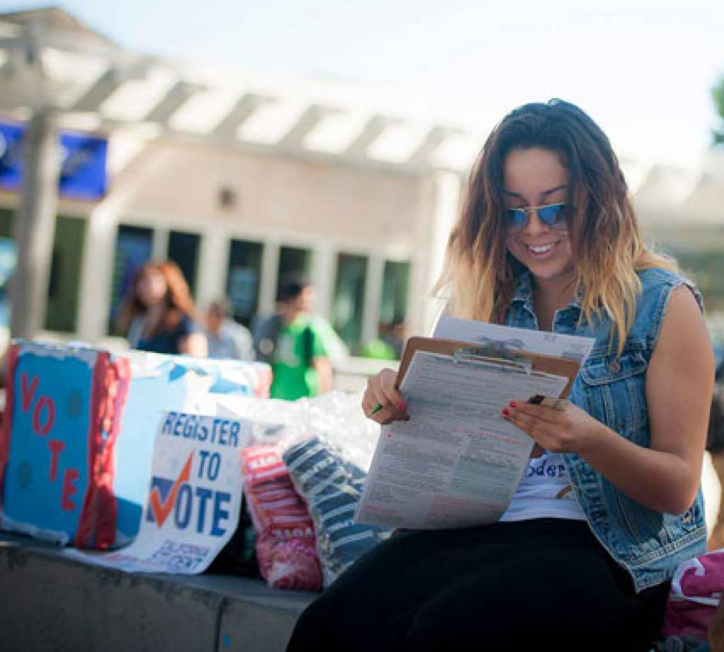 a female student filling out a voter registration form on a clipboard next to "Register to Vote" signs