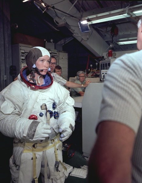 Anna Lee Fisher, wearing a spacesuit with no helmet, with a businesslike expression in a room full of men.