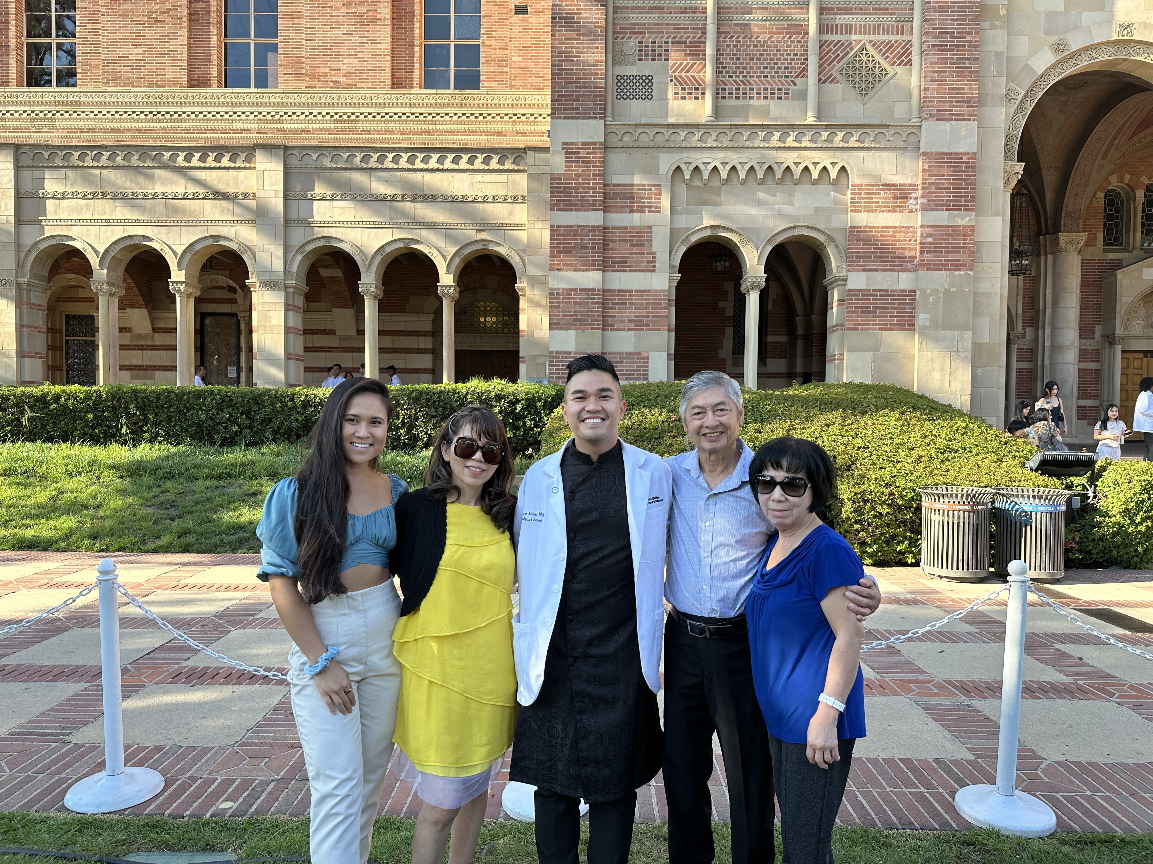 A family of five smiles for the camera with arms around each others' shoulders in front of a stately brick building. An adult son in the center wears a white doctor's coat.