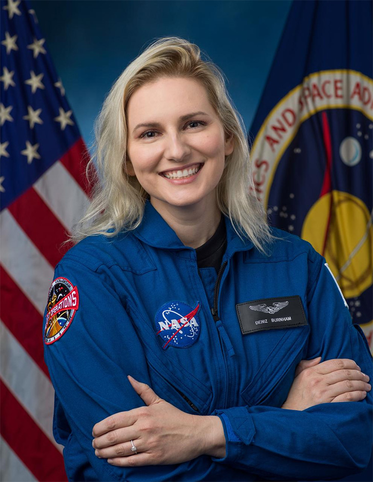 Deniz Burnham official astronaut candidate portrait - posing with arms folded over chest in a blue jumpsuit with American and NASA flags in the background