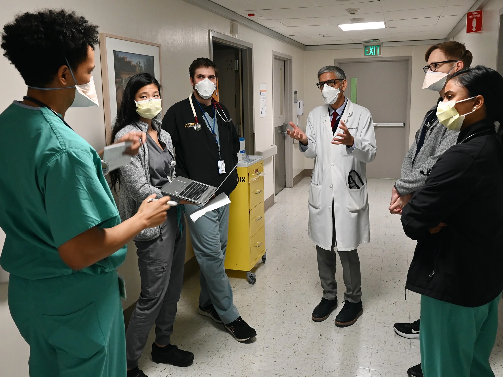 Male doctor in a white coat speaks to a group of medical students wearing scrubs in a hallway of a hospital