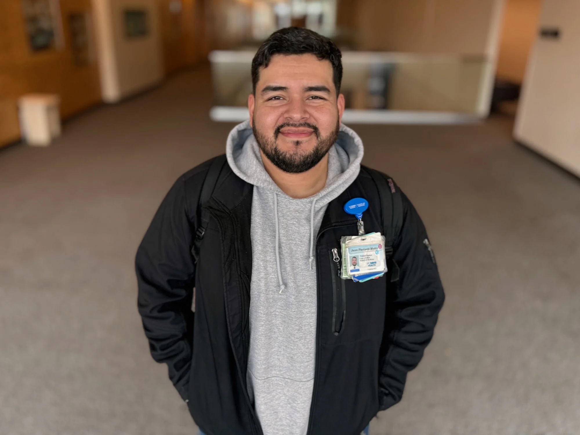 Juan Florio Muniz, young Latino man with a trim beard wearing a gray hoodie and black jacket, smiles at camera with his hands in his jacket pockets