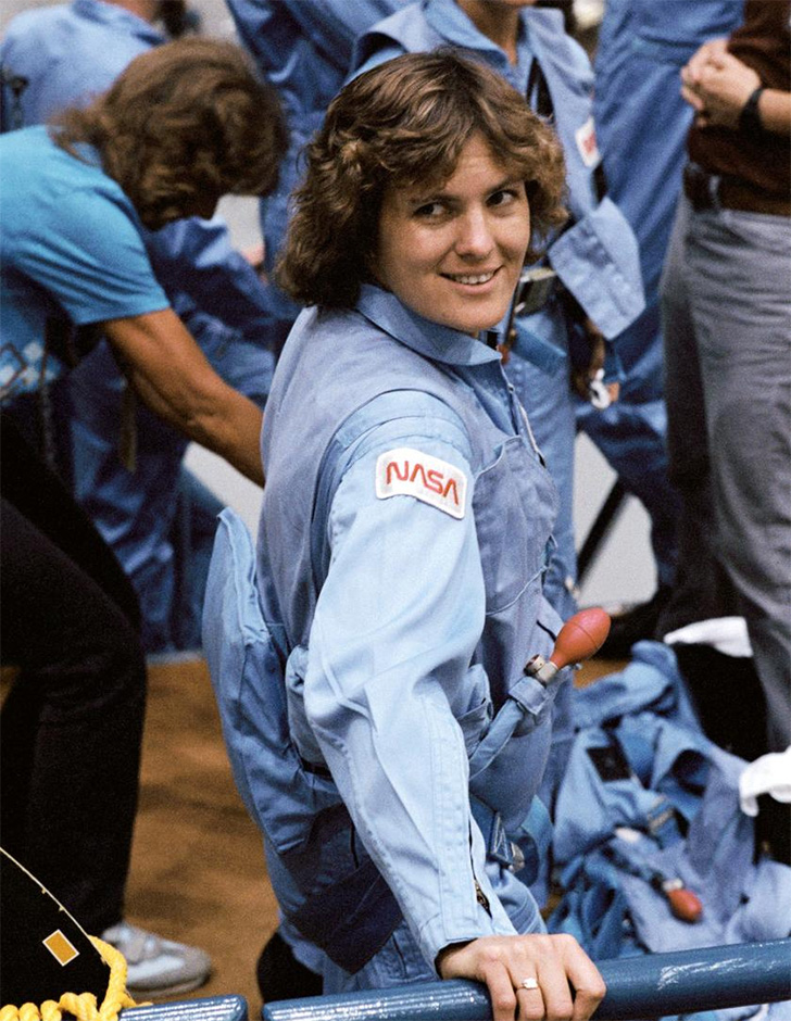 Kathryn Sullivan, with a cool 80's shag haircut, wearing a blue NASA jumpsuit, looks back over her shoulder and smiles