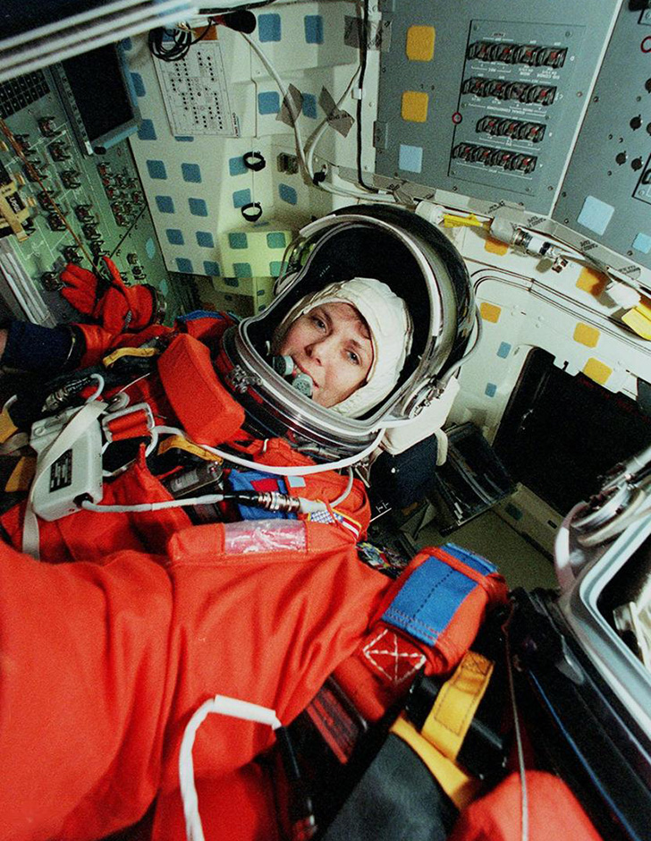 Mary Weber in an orange spacesuit in front of a panel with many switches and instruments