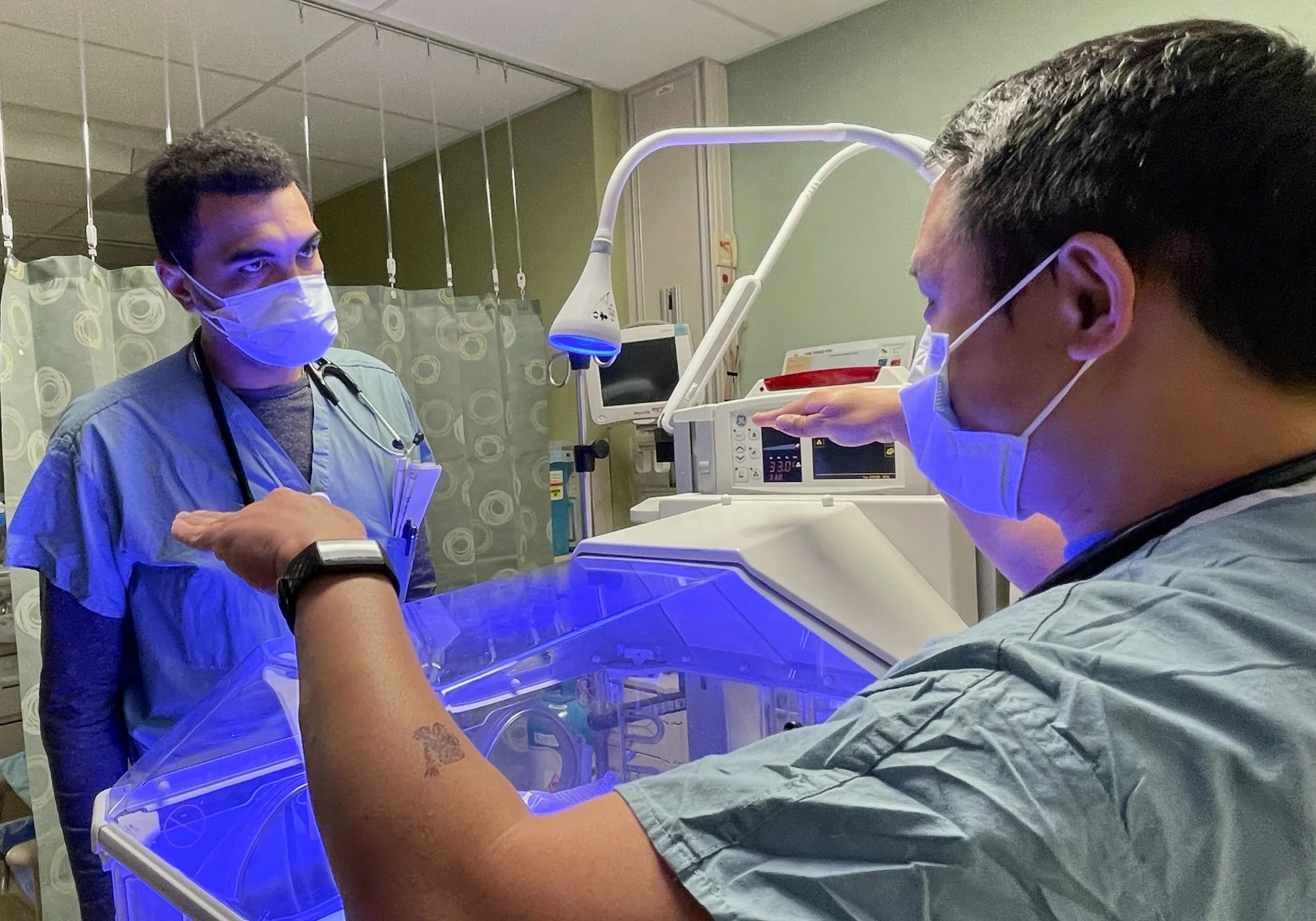 Two men in scrubs and face masks work in a hospital ward, lit with blue light from an instrument