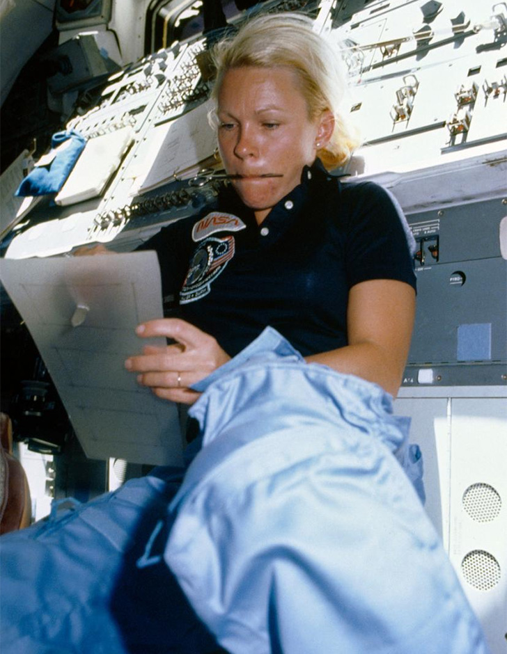 Rhea Seddon uses a sharp tool to punch through a piece of plastic while holding a pen in her mouth in zero gravity