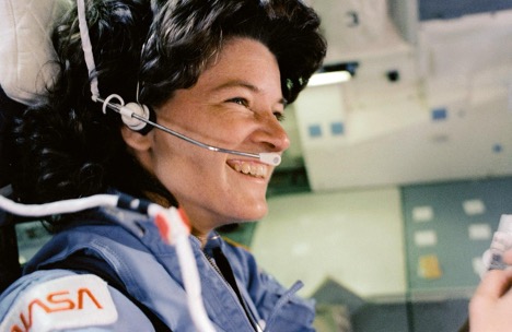 Sally Ride, wearing a headset microphone and a NASA suit, smiles broadly while focusing on a technical task