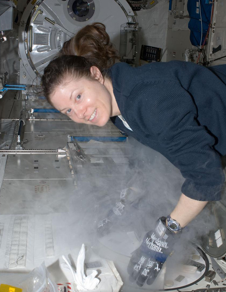 Tracy Caldwell Dyson wears big gloves and smiles at the camera, pausing while working with a cryogenic tank aboard the International Space Station