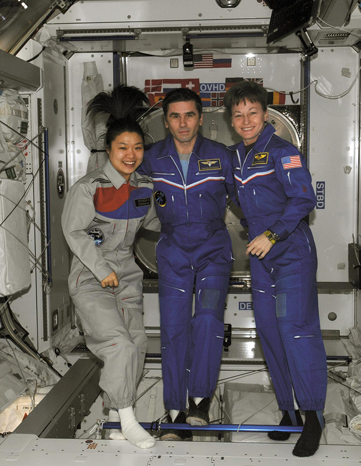 Yi So-Yeon in a gray jumpsuit smiles with two people in blue jumpsuits in a space station in zero gravity